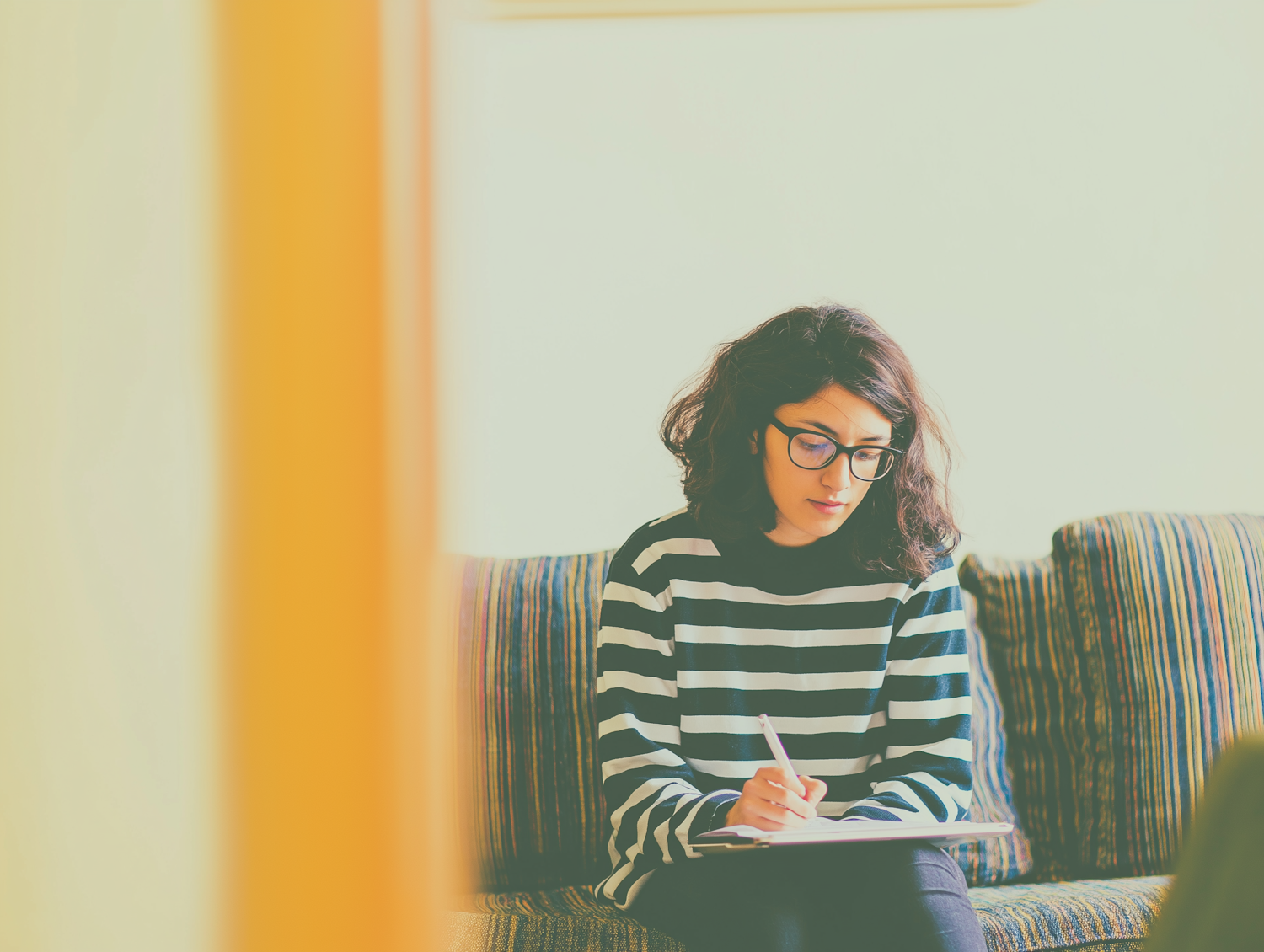 A woman in a striped jumper sitting on a striped sofa, writing in an open notebook in warm indoor light